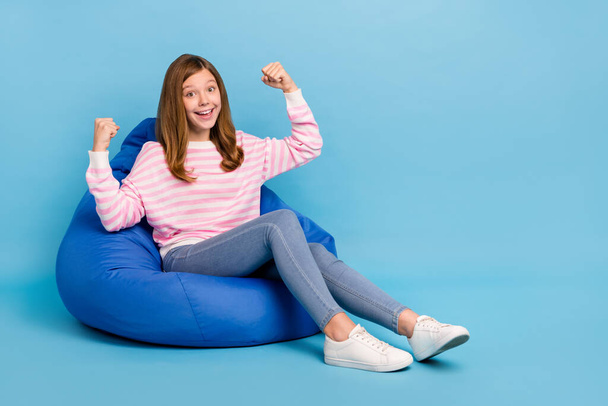 Full size portrait of delighted positive girl sit big soft chair raise fists success isolated on blue color background. - Photo, image