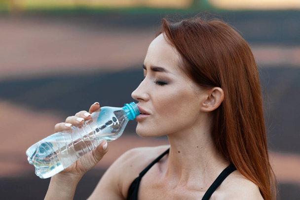 An adult woman drinks water from a bottle after a workout. Portrait of a strong sportswoman. - Foto, imagen