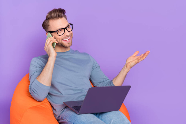 Photo of dreamy funny guy wear grey shirt arm spectacles communicating device empty space sitting beanbag isolated violet color background. - Zdjęcie, obraz