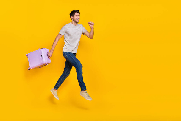 Full size photo of nice brunet millennial guy run with bag wear t-shirt jeans footwear isolated on yellow background. - 写真・画像