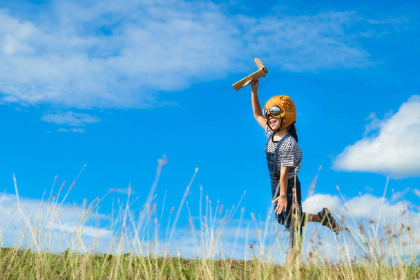 Słodka dziewczynka biegnie przez łąkę w słoneczny dzień z zabawkowym samolotem w ręku. Szczęśliwy dzieciak bawiący się kartonowym samolotem na tle błękitnego letniego nieba. Pomysł wyobraźni z dzieciństwa. - Zdjęcie, obraz