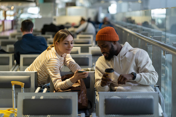 Piękna Europejka prosi przystojnego Afroamerykanina o pomoc w podłączeniu wifi. Młoda para ogląda razem wideo lub zdjęcia w mediach społecznościowych przez telefon komórkowy. Nowi znajomi podczas opóźnienia lotu. - Zdjęcie, obraz