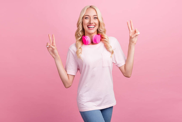 Fotografie sladké tisícileté blond dáma show v-sign nosit sluchátka tričko izolované na růžové barevné pozadí. - Fotografie, Obrázek