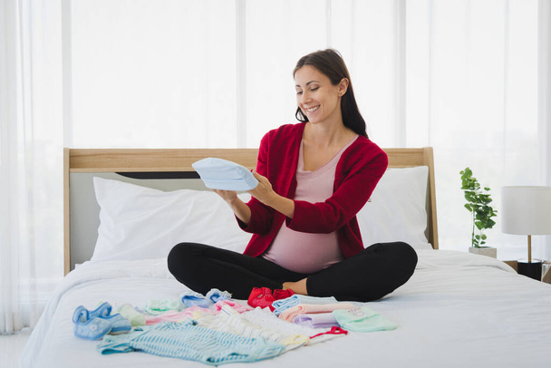 Έγκυος γυναίκα σε casual outfit σχέδιο και να προετοιμάσει τα παιδιά του / της στολή για το μέλλον τους στη σύγχρονη κρεβατοκάμαρα. - Φωτογραφία, εικόνα