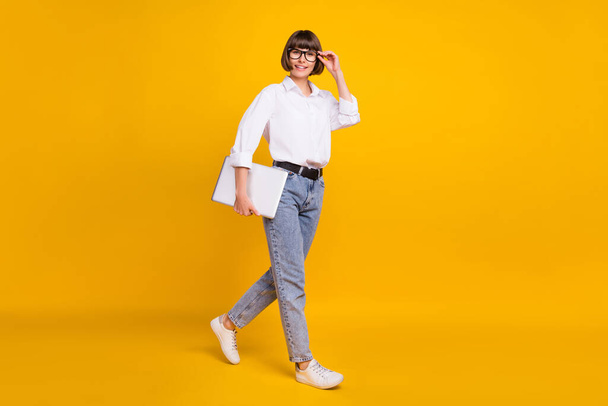 Photo of confident lady carry laptop go wear eyeglasses white shirt jeans sneakers isolated yellow color background. - Foto, afbeelding