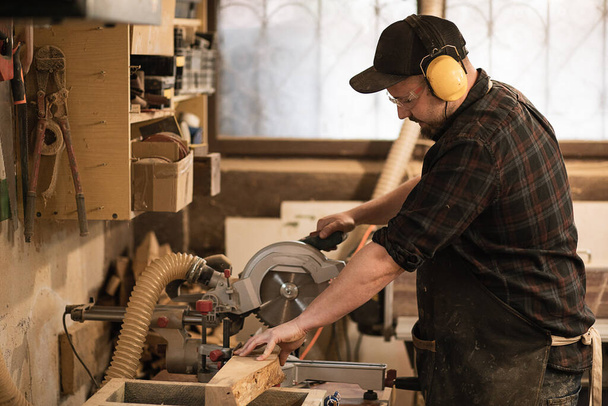 Puuseppä työntekijä suojaava esiliina ja kuulokkeet käyttää modernia hiomakone käsitellä leikkaus puulevyt, puutavaran kanssa pyörösaha työpaja, puutyö ammatti. Pienyritys. Rakennustalo - Valokuva, kuva