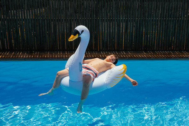 インフレータブルスワンマットレスで10代の男の子日光浴、子供はスペインの夏の間に透明な水でスイミングプールで休息し、リラックスします。水平 - 写真・画像