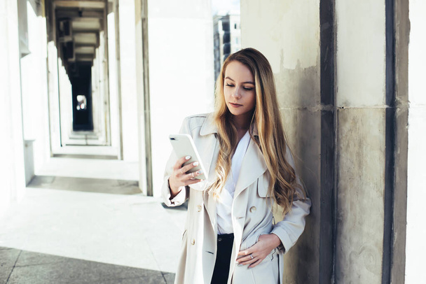 Концентрована молода студентка з довгим світлим волоссям у стильному пальто, що стоїть у довгому коридорі, а текстові повідомлення через смартфон
 - Фото, зображення