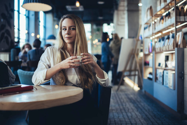 Young female with long hair and mug of coffee looking at camera at table on blurred background of cafe interior - Photo, image