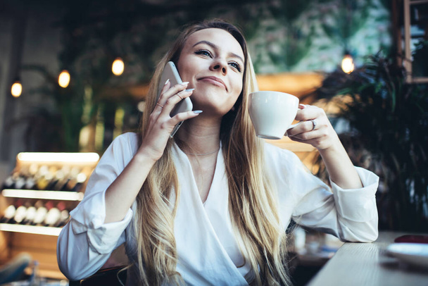 Low angle of cheerful young blond long haired female in white blouse having conversation on mobile phone and drinking hot coffee in cafe - Zdjęcie, obraz