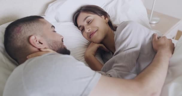 Loving couple lying in bed kissing each other good morning. Happy husband and wife cuddling in bed together in their modern bedroom. Young married couple waking up together in their new home. - Materiał filmowy, wideo