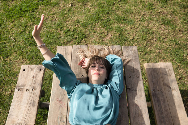Portret van een jonge, mooie, blonde vrouw in groen shirt, die de zonnestralen blokkeert met haar hand, liggend op een houten tafel. Concept warmte, zon, huidkanker, lente, zomer. - Foto, afbeelding