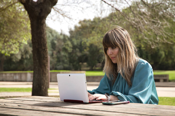 Молода, красива блондинка в зеленій сорочці, сидить на дерев'яній лавці в парку, працює зі своїм ноутбуком. Технологія концепції, планшет, ноутбук, телекомунікації, телекомунікації
. - Фото, зображення