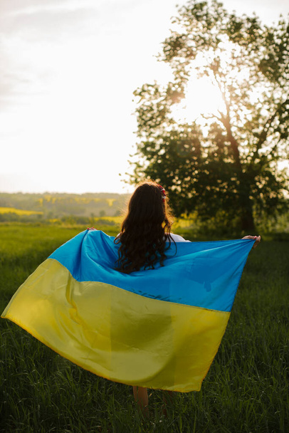 刺繍されたウクライナの伝統的なドレスのウクライナの女性は、フィールド内のウクライナの大規模なフラグを持つフィールドに彼女の背中で立っています.ウクライナのために祈る. - 写真・画像