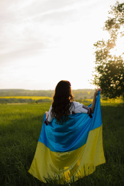 Eine Ukrainerin in bestickter ukrainischer Tracht steht mit dem Rücken auf einem Feld, auf dem eine große Fahne der Ukraine weht. Betet für die Ukraine. - Foto, Bild