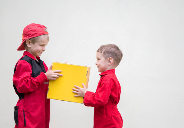 Két cuki, piros overálos kisfiú passzol kézről kézre sárga kartondobozra, könnyű háttérrel. Küldemény, kis postás. Dolgozz úgy, mint egy apa. hirdetési szöveg helye. humoros gyors szállítás hirdetés - Fotó, kép