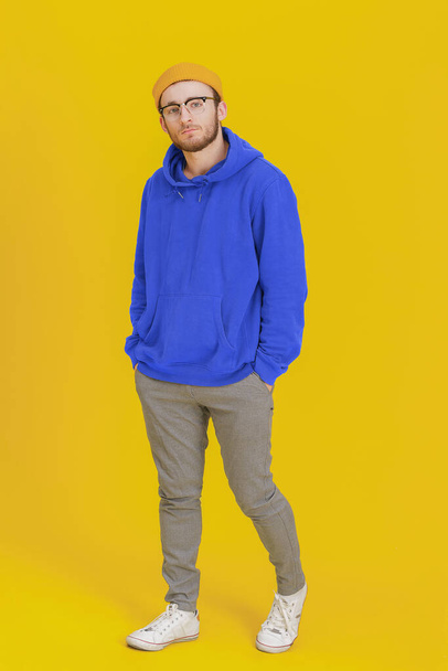 Young man model in casual wear, glasses posing looking at camera on yellow background with hands in pockets. Stylish bearded smart hipster man casual look. Thoughtful guy on yellow background.  - Фото, изображение