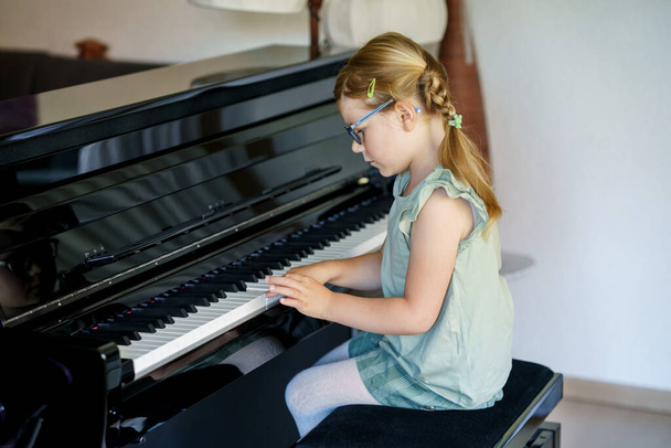 Beautiful little preschool girl playing piano in living room. Cute preschool child with glasses having fun with learning to play music instrument. Early musical education for children. - Photo, Image
