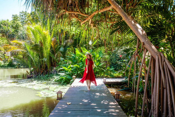 Travel woman in red dress relax in tropical resort. Female traveler on exotic summer vacation. Girl in straw hat walking on wooden bridge near lake in garden with palm trees and green plants - Photo, image