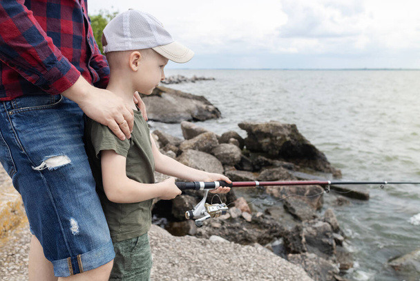 父と息子の釣り。父は息子に回転を保持し、リールを回転させる方法を示します。池や川での釣りの訓練。親の考え方. - 写真・画像