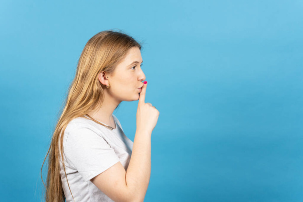 Seitenansicht der jungen blonden Frau bittet um Stille Finger auf Lippen isoliert auf blauem Hintergrund. - Foto, Bild