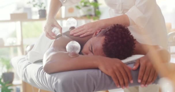 Young mixed race man enjoying relaxing and therapeutic back massage treatment with a vacuum cup in a spa. Beauty therapist treating body pain with suction cupping therapy to remove toxins. - Materiaali, video