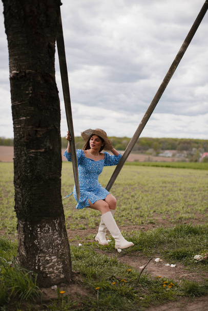 Ένα κορίτσι με μακριά σκούρα μαλλιά, με μπλε φόρεμα, λευκές μπότες και ψάθινο καπέλο, καβαλάει μια κούνια στη φύση.. - Φωτογραφία, εικόνα