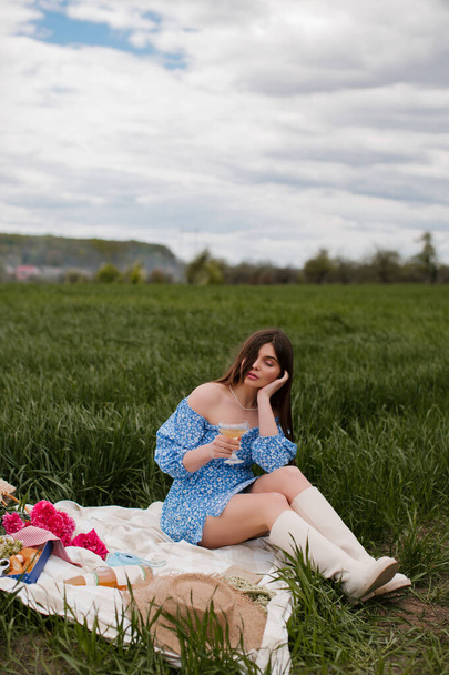 Een mooie jonge vrouw in een blauwe jurk met make-up en roze bloemen drinkt wijn tijdens een picknick in de natuur. - Foto, afbeelding