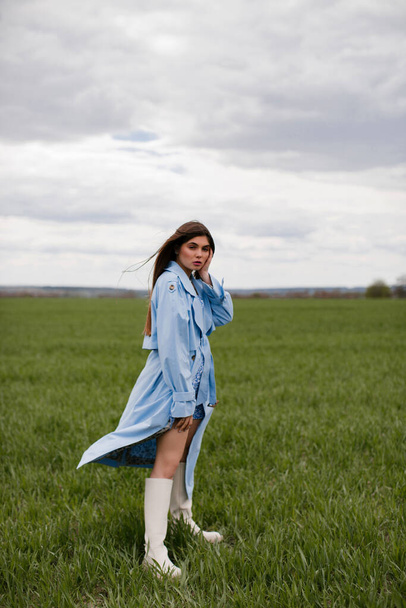 Stylish young woman with makeup and long dark hair posing in a blue trench coat in a green field. - Fotografie, Obrázek