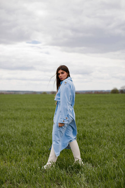Stylish young woman with makeup and long dark hair posing in a blue trench coat in a green field. - Foto, Imagem