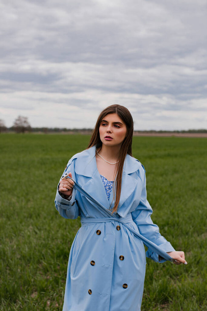 Stijlvol meisje met lang donker haar poserend in een blauwe trench jas in een groen veld. Portret van een jonge vrouw in de natuur. - Foto, afbeelding