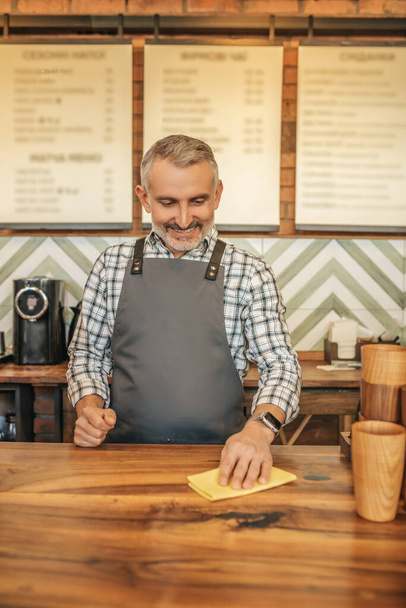 Accuracy. Cheerful gray-haired man in apron smiling looking at surface of bar counter wiping with napkin in cafe - Foto, imagen