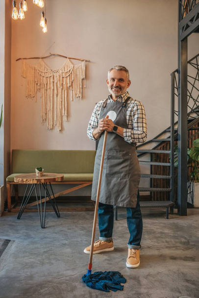 Floor cleaning. Gray-haired joyful middle-aged man in apron holding mop standing smiling at camera in cafe - Foto, Bild