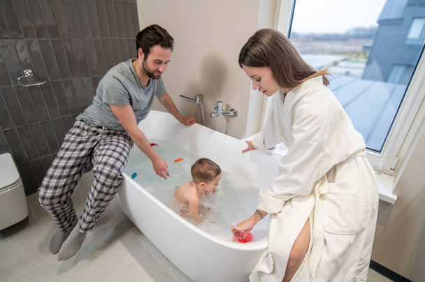Parents, care. Young attentive woman and man in home clothes sitting on bathroom bathing little cute baby - Photo, Image