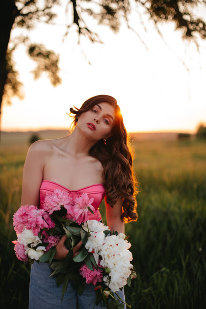 Stylish girl with long wavy hair holds a bouquet of peonies in the field at sunset. - Photo, Image