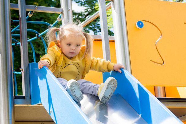 mała dziewczynka w żółtym swetrze sportowym schodzi ze zjeżdżalni na plac zabaw. - Zdjęcie, obraz