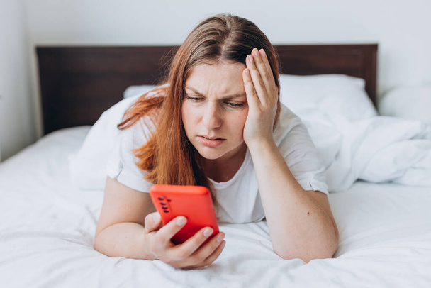 Upset young woman lying in bed feel sick or distressed using modern cellphone gadget, sad millennial female relax in bedroom hold smartphone read bad news - Foto, Imagen