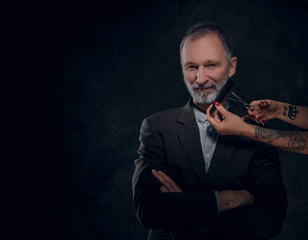 Shot van vrouwelijke handen snijden baard van senior man gekleed in elegante pak tegen donkere achtergrond. - Foto, afbeelding