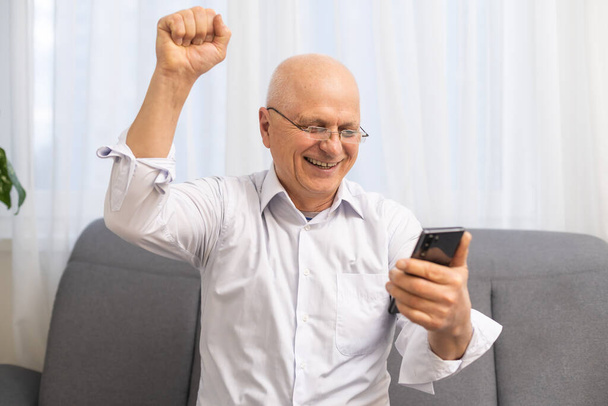 Happy innoissaan isoisä tilalla älypuhelin, nauraa, huutaa iloa, saada hyviä uutisia, lukeminen tekstiviestin ruudulla, jolloin voittaja kyllä käsi ele. Yllättynyt vanhempi mies voittaa palkinnon - Valokuva, kuva