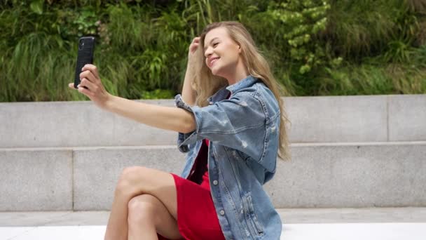 Vista lateral de ángulo bajo de la joven mujer en chaqueta de mezclilla y falda tomando selfie en el teléfono inteligente mientras está sentado en el banco de piedra en la ciudad - Imágenes, Vídeo