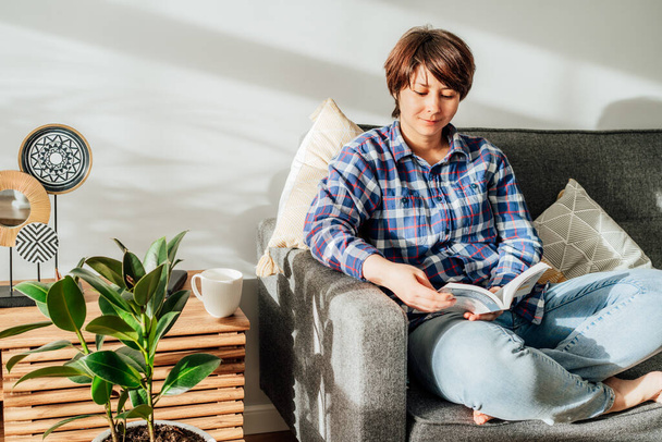 Woman sit on the sofa couch and relax while reading a book at home in cozy modern interior living room with green plants. Offline activity lifestyle. Reading hobby. Leisure indoors entertainment. - Foto, afbeelding