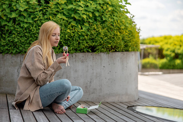 Young beautiful blond woman eating sushi outdoors, on the wooden terrace, by modern building in the city. Tasty food to go. Girl has lunch break, spending time outside, eating Asian food. City life - Photo, Image