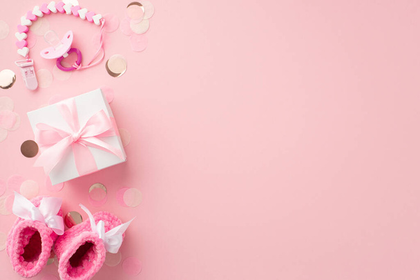 Concept d'accessoires bébé. Photo de vue du dessus de boîte cadeau blanche avec chaîne de sucette papillon rose chaussons tricotés et confettis brillants sur fond rose pastel isolé avec copyspace - Photo, image