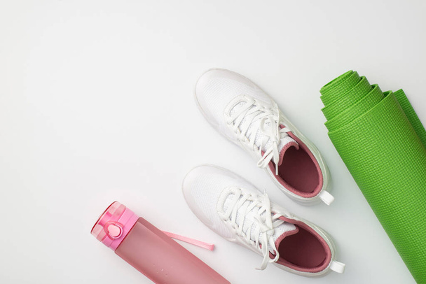 Spor aksesuarları konsepti. Beyaz spor ayakkabıların, pembe su şişesinin ve izole edilmiş beyaz arka planda yeşil yoga minderinin en üst görüntüsü. - Fotoğraf, Görsel