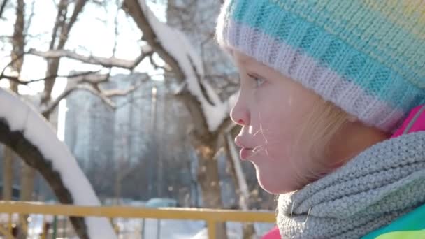 Primer plano de una niña de ojos azules al aire libre en el helado día de invierno. Niño con ropa de abrigo soplando en sus manoplas y puede ver su aliento - Metraje, vídeo