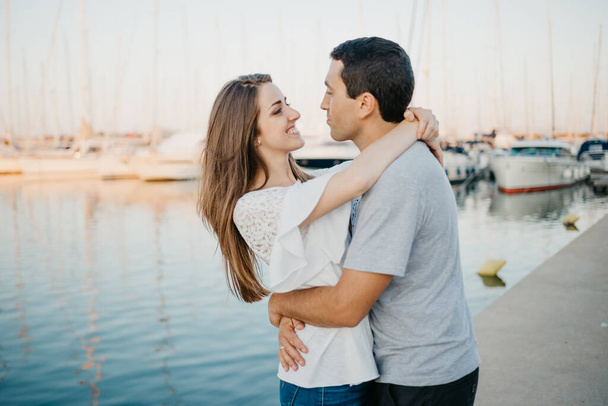 Un homme hispanique étreint sa petite amie brune heureuse au port en Espagne dans la soirée. Un couple de touristes fréquentent près de yachts au coucher du soleil à Valence. - Photo, image