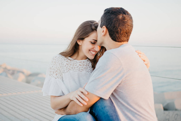 A Hispanic man is kissing on the forehead his smiling brunette girlfriend on a breakwater in Spain in the evening. A couple of tourists on a date at the sunset in Valencia. - 写真・画像