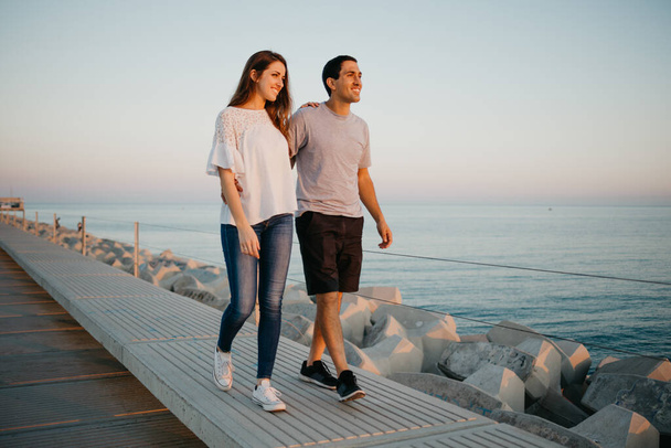 A photo in the full length of a Hispanic man with his brunette girlfriend who are walking on a breakwater in Spain in the evening. A couple of tourists are staring at the sea on a date in Valencia. - Fotó, kép