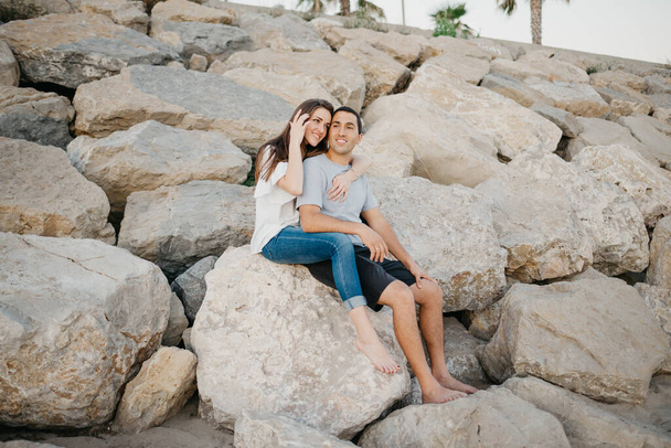 Un hombre hispano está sentado entre las piernas de su novia morena sonriente en las piedras de un rompeolas en España por la noche. Un par de turistas en una cita al atardecer en Valencia. - Foto, imagen