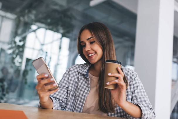 European girl using mobile phone and drinking coffee at desk. Concept of modern successful woman. Young smiling female person wearing casual clothes. Notebook on wooden table - Foto, afbeelding